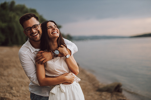 Beautiful wedding couple in shallow water of river in backlight on sunny summer afternoon, groom carrying bride, both smiling at camera