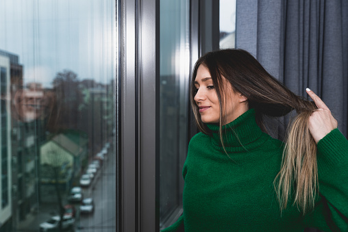 Young happy college student woman looking through the window of her new rented modern apartment feeling exited and relaxed because she now live alone. Female taking hair band of her hair