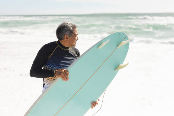 istock Biracial senior man carrying blue surfboard on shore while looking at sea during sunny day 1365871688