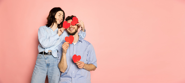 Young and happy man and woman holding greeting cards shaped hearts isolated on pink trendy color background. Human emotions, youth, love and lifestyle concept. Valentine's day celebration. Flyer