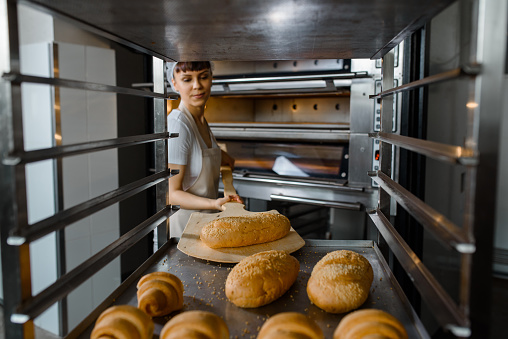 Close up of young happy smiling caucasian woman baker putting the fresh bread on the shelves/rack at baking manufacture factory. Tasty bread bakery factory concept.