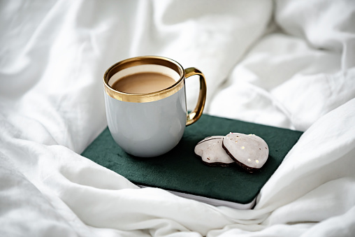 Cup of aromatic coffee, notebook and cookies on white bed linen. Cozy morning at home while reading and having breakfast in bed. Hygge and relaxation concept