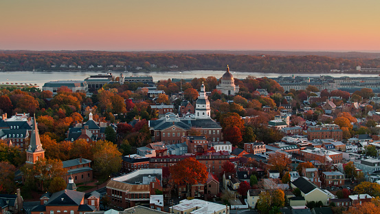 Aerial shot of Annapolis at sunrise on a hazy morning in Fall, looking past the dome of the Maryland State House towards the campus of the US Naval Academy, with Chesapeake Bay beyond.