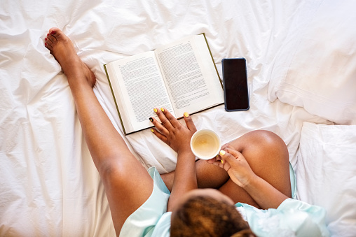 Overhead view of a woman sitting in her bed in the morning with a cup of coffee and reading a book