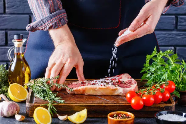 male chef is sprinkling salt over a porterhouse beef steak on restaurant kitchen. man cooking beefsteak