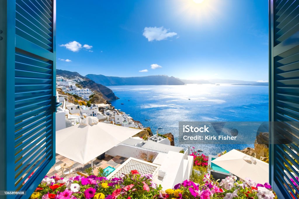 Santorini Window View with Flowers View from an open window with blue shutters of the Aegean sea, caldera, coastline and whitewashed town of Oia, Santorini, Greece. Santorini Stock Photo