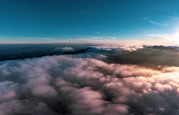 Blue sky above thick layer of white fluffy clouds at bright orange-colored sunrise with mountain peaks underneath panorama view