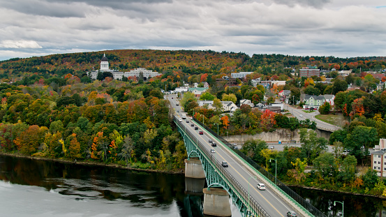 Aerial shot of Augusta, Maine on an overcast day in Fall. The drone flies over the Kennebec River away from Memorial Bridge. The Maine State House can be seen in the distance against a background of trees showing Fall colors.