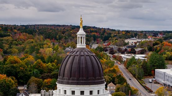 Aerial shot of the Maine State House in Augusta on an overcast day in Fall, with a vibrant autumnal forest behind.