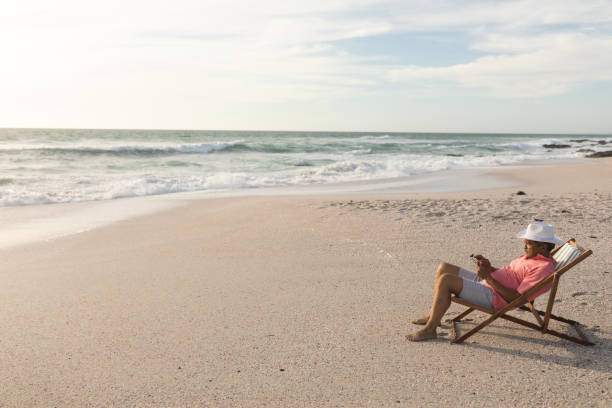 istock Full length side view of biracial senior man using digital tablet relaxing on folding chair at beach 1365904998