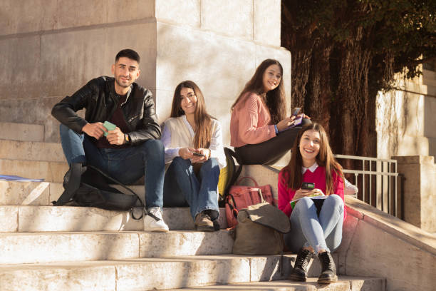 empat siswa melihat kamera dan tersenyum sambil memegang smartphone mereka duduk di tangga universitas. - study abroad potret stok, foto, & gambar bebas royalti