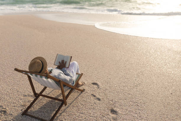 istock Relaxed senior biracial woman reading book while sitting on folding chair at beach during sunset 1365907683