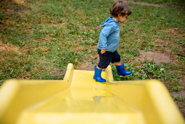 View from behind of two children wearing bright clothing, standing on a metal barrier at a sporting event. They have climbed onto the bottom bar to be able to get a better view. The young boy is wearing blue shorts, yellow socks and grey and red sports shoes. The young girl is wearing colourful shorts with a fruit pattern and a colourful jacket and pink and purple rubber boots.