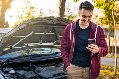 One happy young adult caucasian man standing by his vehicle with open hood and broken failed engine smiling carefree hold phone calling towing service for help on the road Roadside assistance concept