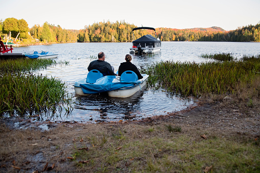 Senior man in a pedal boat heading to his deck boat in autumn. He is with an adult young woman. Horizontal full length outdoors shot with copy space.