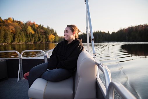 Portrait of healthy young woman enjoying life on a boat in autumn. She is looking at the camera with a smile and is wearing a black hoodie. Horizontal full length outdoors shot with copy space.