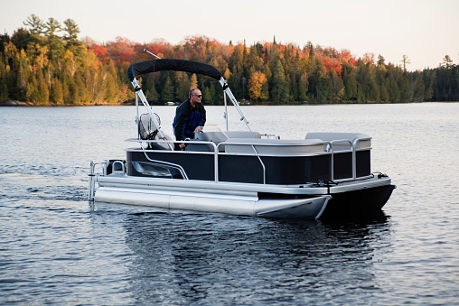 Portrait of happy senior man driving a boat in autumn. He is looking at the horizon with a smile and is wearing warm clothes. Horizontal full length outdoors shot with copy space.