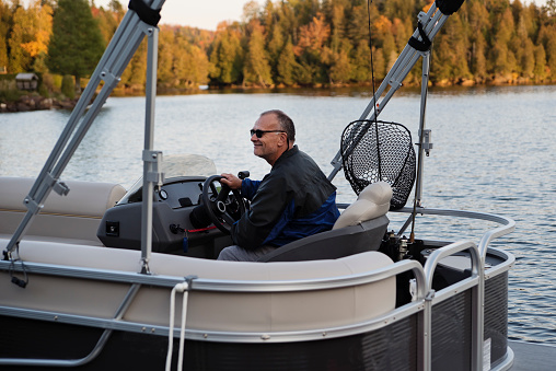 Portrait of happy senior man driving a boat in autumn. He is looking at the horizon with a smile and is wearing warm clothes. Horizontal full length outdoors shot with copy space.