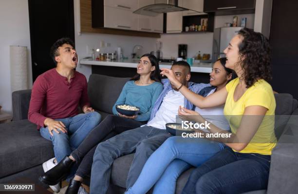 Happy Group Of Flat Mates Eating Popcorn While Watching Tv At Home Stock Photo - Download Image Now