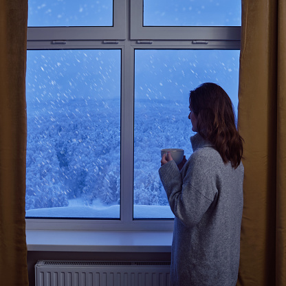 A woman stands at a winter evening window with trees in the snow. A woman with a cup of coffee looks out the window with a snowfall behind the glass