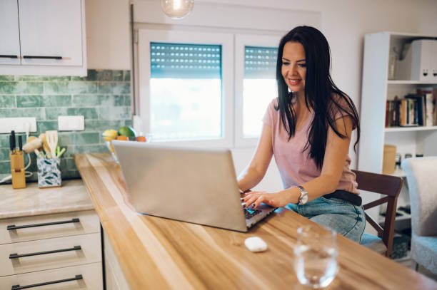 istock Middle aged woman using laptop while sitting at kitchen counter at home 1365940951