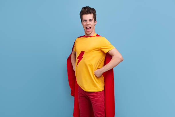 Funny courageous male in bright superhero cape standing with hands on waist and demonstrating strength while looking at camera on blue background in studio