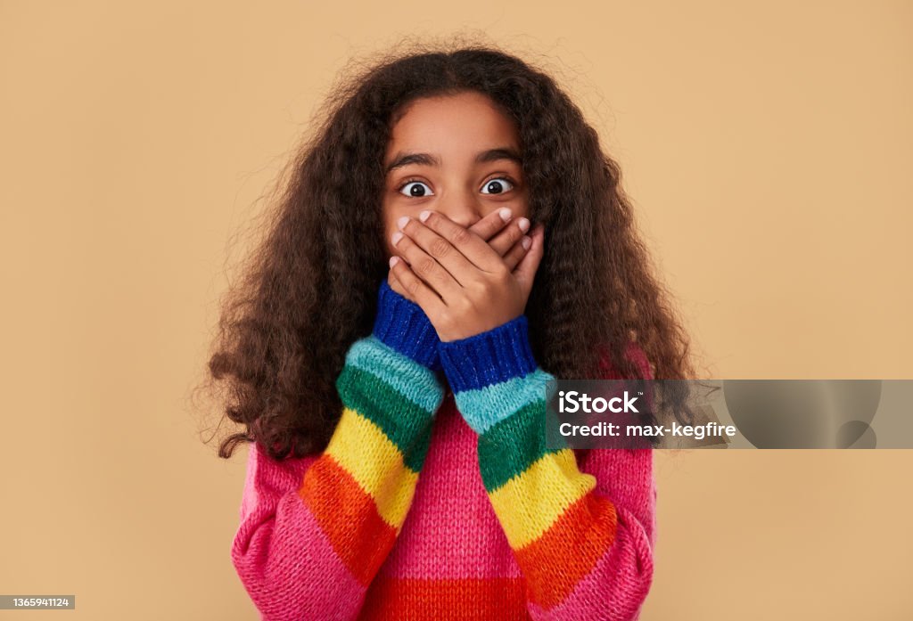 Amazed ethnic girl covering mouth with hands Excited shocked African American girl in colorful knitted sweater covering mouth with hands while keeping secret and looking at camera against beige background Mistake Stock Photo