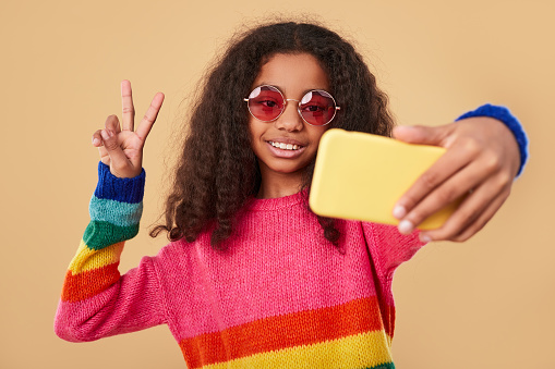 Optimistic teen African American girl with curly hair wearing colorful striped sweater and trendy sunglasses making peace gesture and taking selfie, on smartphone against beige background