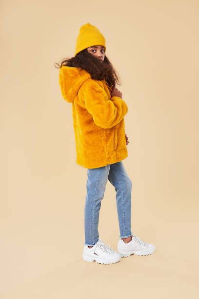 Stylish hipster girl in warm winter outfit Full body side view of African American hipster girl wearing bright yellow warm jacket with knitted hat and jeans standing against beige background coat jacket winter isolated stock pictures, royalty-free photos & images