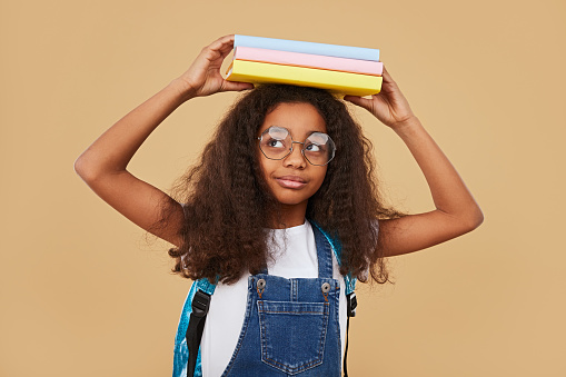 Clever African American schoolgirl with curly hair wearing denim overall and glasses carrying stack of colorful textbooks on head against beige background