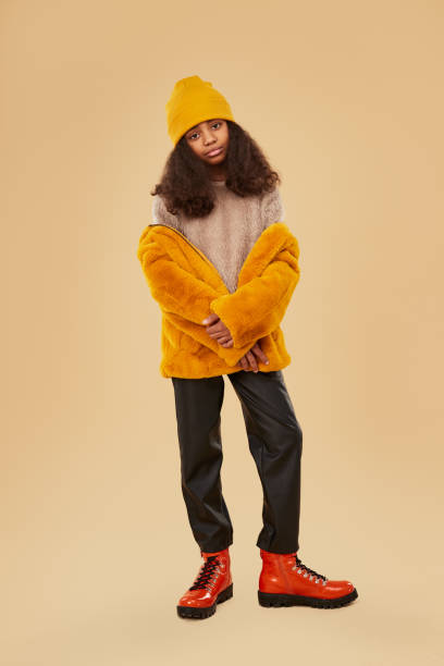 Black teen girl in colorful warm wear Full body of African American teenage girl in bright yellow fur jacket and hat and stylish red boots looking at camera against beige background kids winter fashion stock pictures, royalty-free photos & images