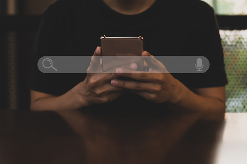 Joven asiático con camiseta negra sentado en el escritorio sosteniendo el teléfono inteligente a la mano en busca de información. Uso de Search Console con tu sitio web. photo