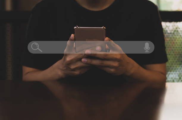 joven asiático con camiseta negra sentado en el escritorio sosteniendo el teléfono inteligente a la mano en busca de información. uso de search console con tu sitio web. - spotting fotografías e imágenes de stock