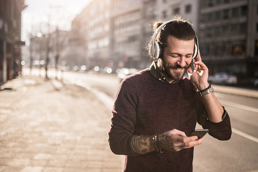 Cheerful young man using smart phone on city street and listening to music on wireless headphones.