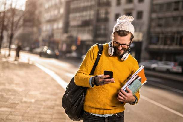 jeune étudiant universitaire marchant dans la rue de la ville et utilisant son téléphone. - headphones relaxation outdoors caucasian photos et images de collection