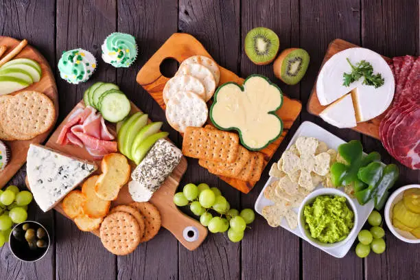 St Patricks Day theme charcuterie table scene against a wood background. Selection of cheese, meat, fruit and vegetable appetizers. Above view.