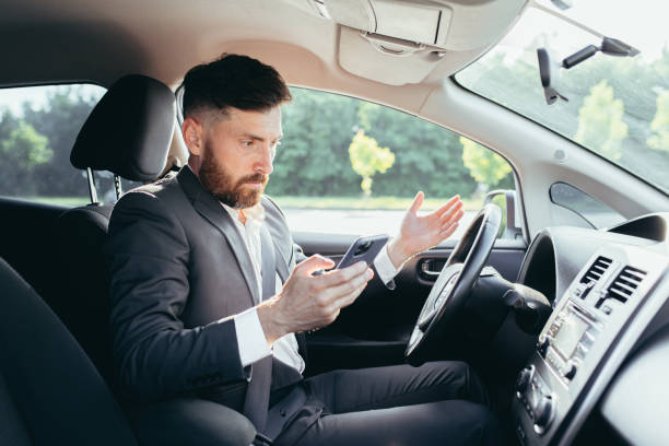 мужчина-бизнесмен, сидящий за рулем автомобиля, расстроенный не может понять рулевое управление и инструкции фанкового автомобиля, читает  - driving mobile phone car talking стоковые фото и изображения