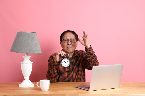 The senior Asian man sitting at working desk with laptop on the pink background.