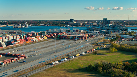 Drone shot of containers stacked on the docks in Portsmouth Marine Terminal in the Port of Virginia, with military ships in berths at the end of the dock and across the river in Norfolk Navy Yard. A line of trucks is waiting to enter the terminal.