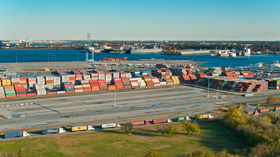 Drone shot of containers stacked on the docks in Portsmouth Marine Terminal in the Port of Virginia, with military ships in berths at the end of the dock and across the river in Norfolk Navy Yard. A line of trucks is waiting to enter the terminal.