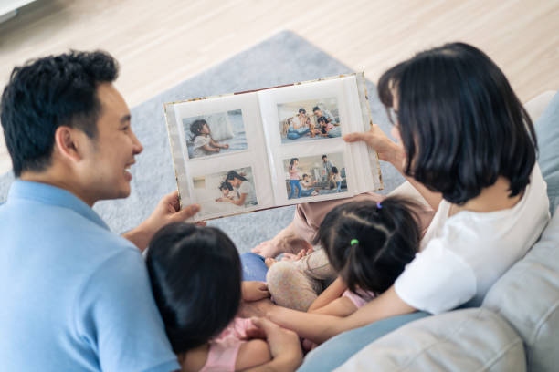 Asian family look at photographs on old picture album recalls the past. Young beautiful couple sitting with little kid daughters flipping pages of photo book and enjoy remembering relationship memory. stock photo