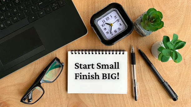 istock Start small finish big text on notebook with glasses, pen, clock, plant, laptop, and brown table background. Business concept. 1365977144