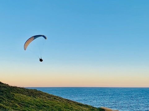 Horizontal seascape of paragliding enthusiasts hovering over grassy headland over ocean at sunset at Lennox Head beach lookout NSW Australia