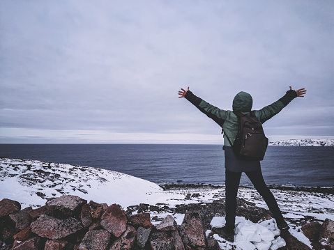Winter, man above the rocky cliff raises his arms up facing the horizon of the sea, concept of freedom. a traveler on the edge of the earth. The Arctic Ocean.
