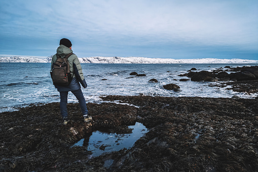 Traveler standing on icy shore near sea. tourist in a warm down jacket with a camera and a backpack is standing on the edge of the earth. The Arctic Ocean.