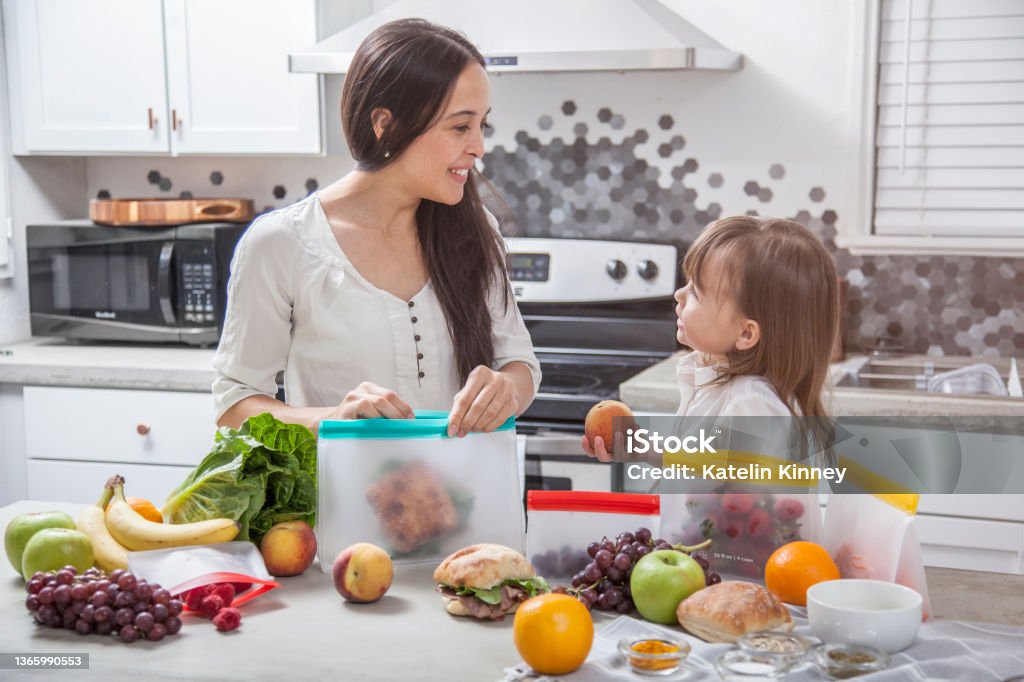 A mother and daughter pack sandwiches and fresh food into clear storage bags in a bright modern kitchen. A young mother puts a sandwich into a clear food storage bag. Her daughter is handing her a peach. They are in a bright modern kitchen with a concrete counter top surrounded by other fruits and vegetables that she is preparing and storing. Healthy Eating Stock Photo