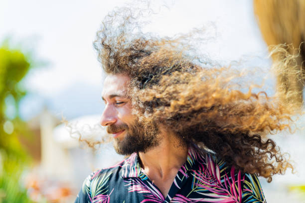 Portrait of happy young sporty man touching his long hair, enjoying freedom after workout by the sea, smiling to camera. Concept of successful outdoor fitness training session