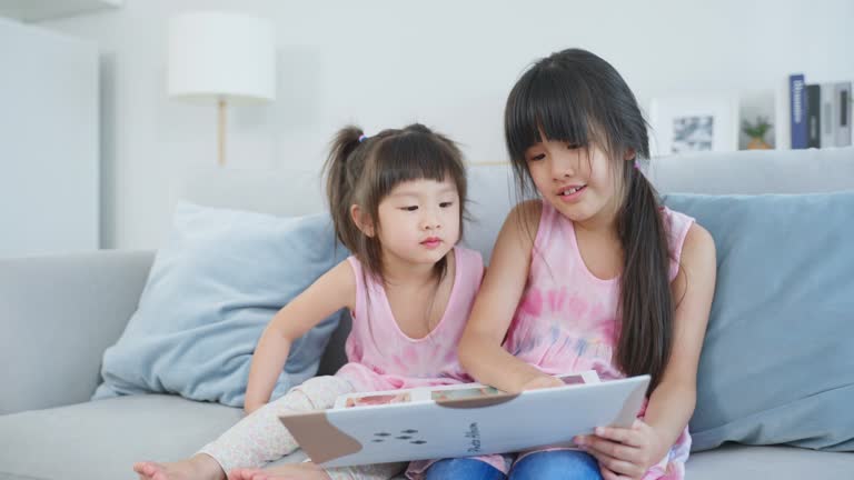 Asian big sister showing old album recalls the past to younger sibling. Young adorable children girl sitting on sofa, flips pages of photo book and enjoy remembering relationship memory in photographs