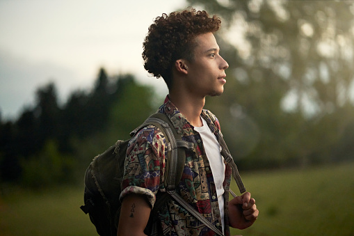 Waist-up profile view of contemplative man with curly brown hair wearing casual clothing, backpack, and looking away from camera at dusk.