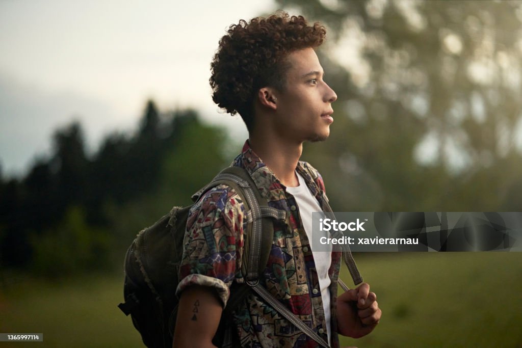 Outdoor portrait of young African-American backpacker Waist-up profile view of contemplative man with curly brown hair wearing casual clothing, backpack, and looking away from camera at dusk. Profile View Stock Photo